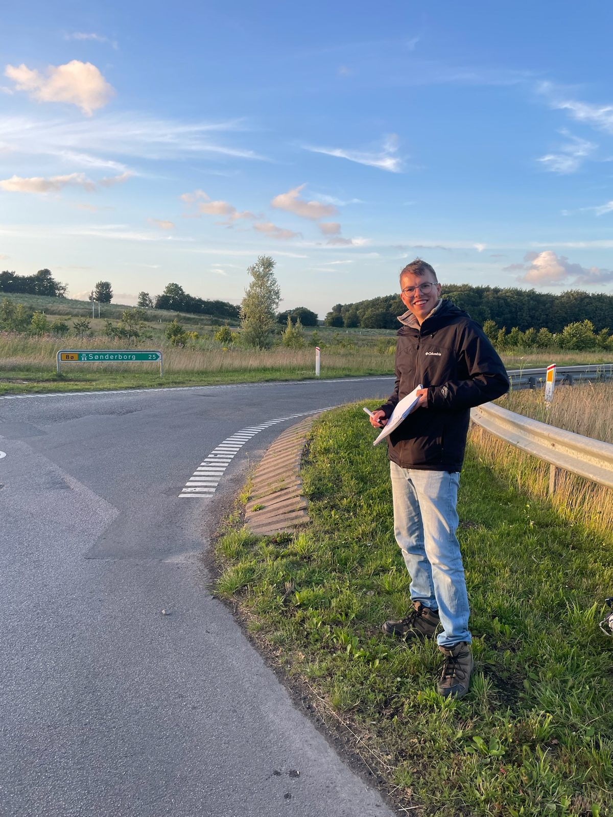 Johannes standing on the grass verge behind a metal barrier near Sønderborg, holding his notebook sign